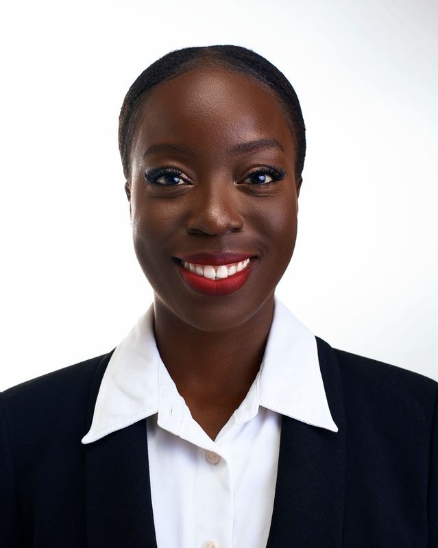 Grace A. Owusu, a woman in a black suit and white collared shirt, smiling against a white backdrop